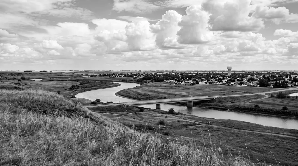 Generated black-and-white scene: a prairie river valley seen from a high grassy bank, with a low road bridge and a small city on the far bank; not a photograph of North Battleford.